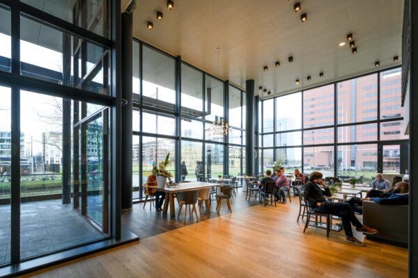 Moderne en ruime ontmoetingsruimte met grote ramen aan de Thomas R. Malthusstraat, waar mensen ontspannen en met elkaar praten.